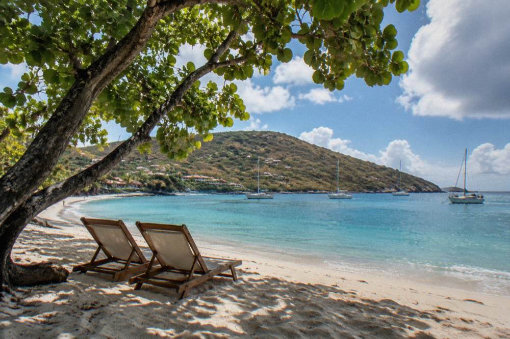 Two beach chairs on pale sand under leaning green trees at a Caribbean bay