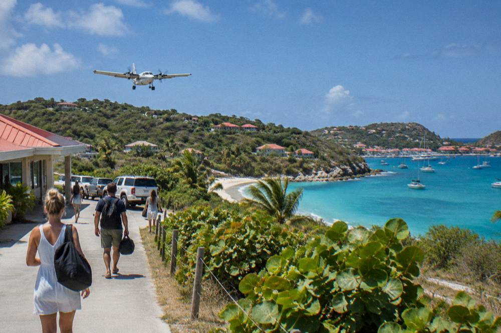 Travellers with small soft bags walking from a tiny airport terminal toward parked taxis