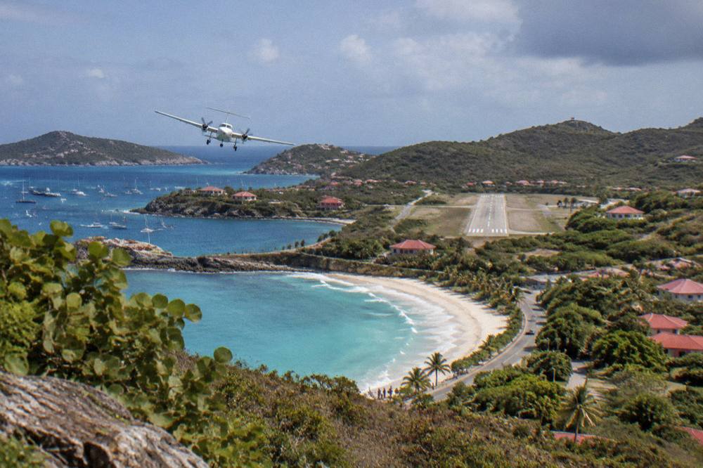 A small plane banking low over a beach road toward a runway between hills