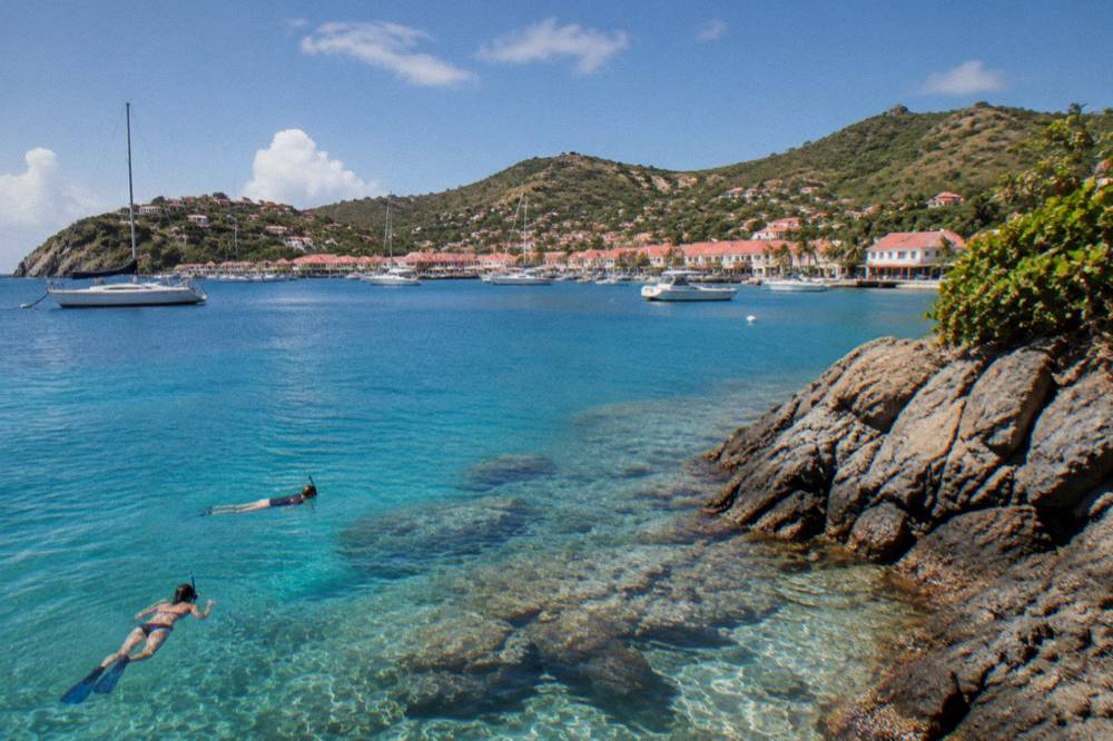 Snorkellers in clear water above rocks and coral near a rocky headland