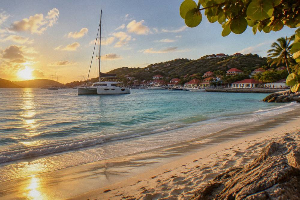 A sailing catamaran anchored in a green bay with a sandy beach and clear water